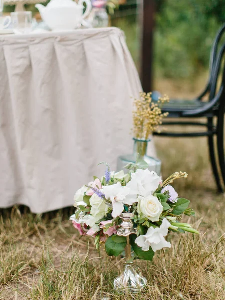 A bouquet of white and lilac eustoma, pink roses, twigs, and leaves stands in a copper candlestick on the grass. In the background is a banquet table with a white tablecloth.