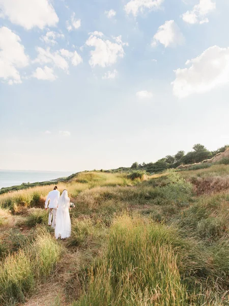 The groom in a beige suit and the bride in a white dress with a long veil is walking along a hill overgrown with steppe grasses. The sea is in the background.
