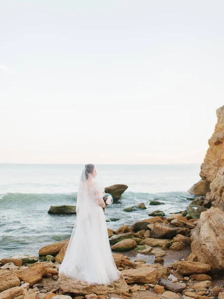 The bride in a white long dress and veil holds a wedding bouquet and stands on a rocky beach. In the background, there is a rock and the sea.