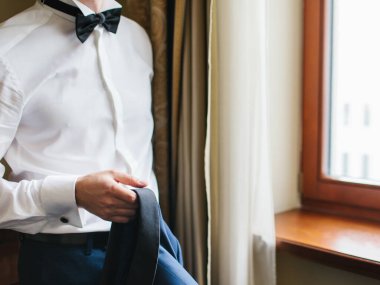 A man is standing by the window in a white shirt and a black bow tie, holding a dark blue jacket in his hand.