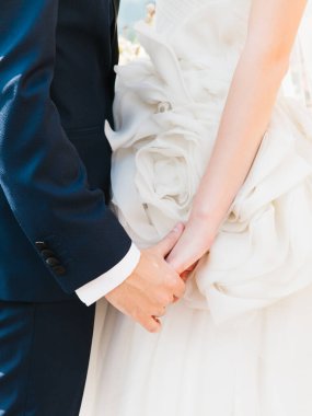 Newlyweds hold hands close-up. The groom is in a dark blue suit, the bride in a white airy dress with flowers.