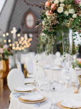 Wedding table setting in gold and white. On the table are a white tablecloth, dishes, cutlery, and a bouquet of flowers in a tall glass vase. In the background is a brown tower with an oval window.