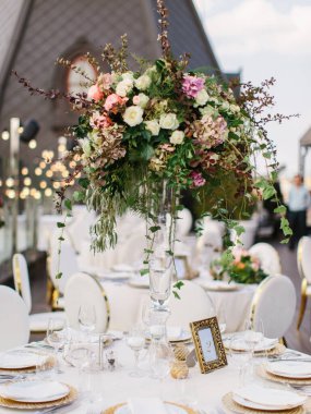 The decor of the wedding banquet is white on the roof against the backdrop of a brown tower with an oval window. On the table are a white tablecloth, cutlery, plates, wine glasses, and bouquets of flowers.