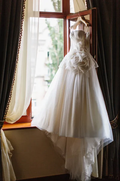 A fluffy wedding dress with a fabric flower and a long train hangs on a hanger in an open wooden window.