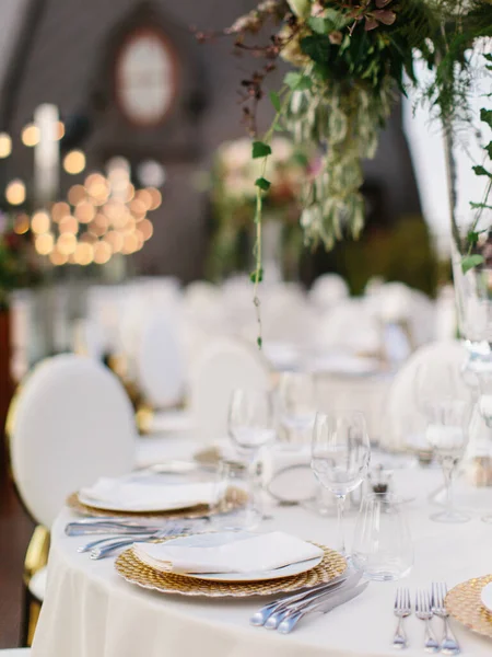 Wedding table setting in gold and white. On the table are a white tablecloth, dishes, cutlery, and a bouquet of flowers in a tall glass vase. In the background is a brown tower with an oval window.