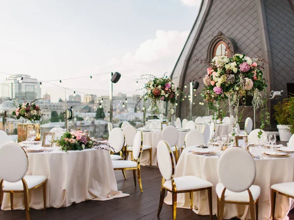 The decor of the wedding banquet is white on the roof against the backdrop of a brown tower with an oval window. On the table are a white tablecloth, cutlery, plates, wine glasses, and bouquets of flowers.