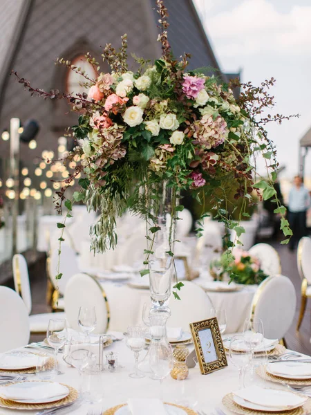 The decor of the wedding banquet is white on the roof against the backdrop of a brown tower with an oval window. On the table are a white tablecloth, cutlery, plates, wine glasses, and bouquets of flowers.