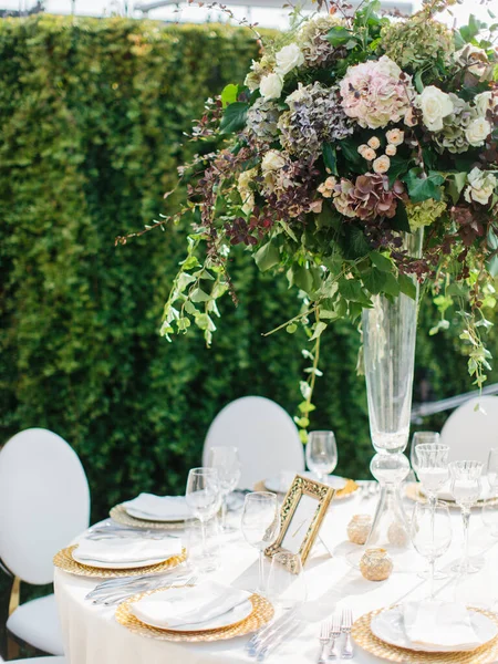 Serving a wedding banquet table in white tones against a background of greenery. On the table is a bouquet in a glass vase of roses and hydrangeas, white and gold plates, wine glasses, and cutlery.