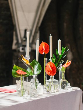Bouquets of red anthurium, strelitzia, and tropical leaves stand in glass vases on a white table. There are white candles in the background. Tropical decor.