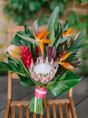 The bride's bouquet in a tropical style with exotic flowers stands on a wooden chair. Bouquet of protea, palm leaves, strelitzia, and red ginger.