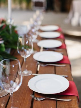 Decor and serving of a wedding banquet on a wooden table.White plates on a red napkin, cutlery, and wine glasses. Tropical flowers in the background.