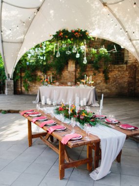 The decor of the wedding banquet is a tropical style in a white pavilion. The wooden table is set with a white tablecloth, exotic flowers, candles, wine glasses, dishes, and red napkins on plates.