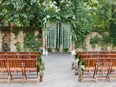 Tropical wedding decor. Rows of wooden chairs decorated with pineapple and palm leaves, wedding arch decorated with leaves, candles, and macrame.