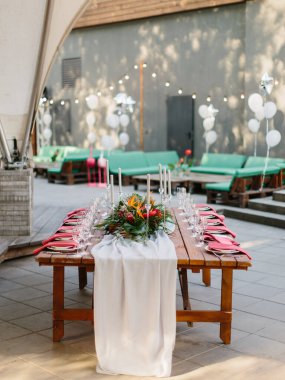 The decor of the wedding banquet is in a tropical style in the pavilion. The wooden table is set with a white tablecloth, exotic flowers, candles, wine glasses, dishes, and red napkins on plates.