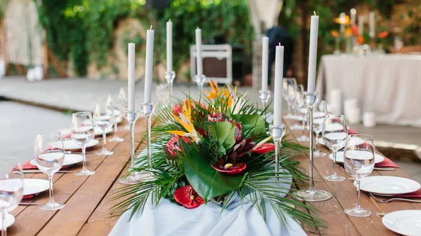Wedding banquet decor in tropical style. In the center of the table is a white tablecloth, a bouquet of strelitzia, red anthurium, and tropical leaves. On the sides are candles, wine glasses, and plates.