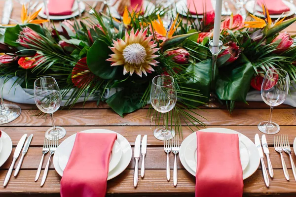 Decor and serving of a wedding banquet in a tropical style. Exotic flowers, protea, strelitzia, leaves, white candles in high candlesticks, wine glasses, red napkins on white plates, cutlery on a wood