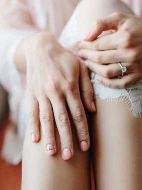 A bride with a nude manicure in an engagement ring with a gemstone keeps her hands on her knees. On the bride's ring finger on the nail, inscribe 