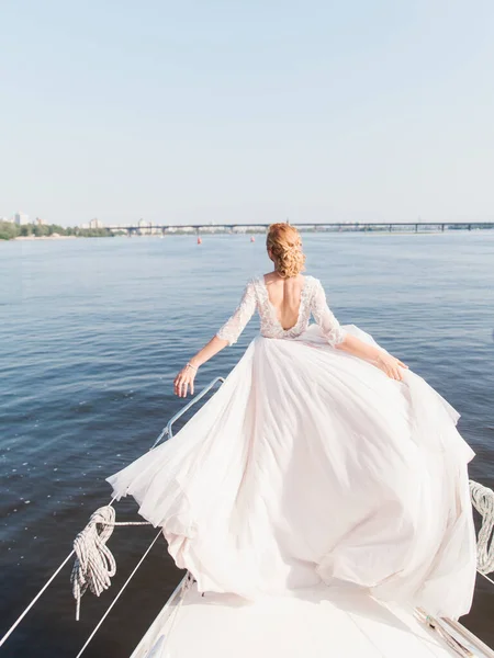 A bride with blond hair in a fluffy white dress with an open back stands on the bow of the yacht with her arms outstretched. The train of the dress is evolving. Ahead is a river landscape and a bridge