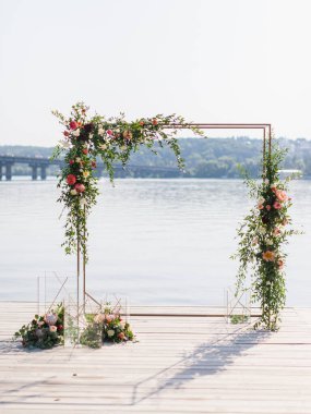 A golden square wedding arch decorated with white, pink, and red peonies stands on a wooden pier. Below are bouquets of flowers and decorative golden cubes. In the background, there is a river.