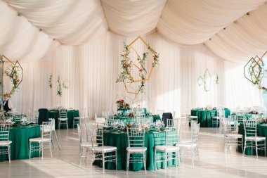 The decor of the wedding banquet hall with tables in emerald green, white drapery on the ceiling, and gold geometric decorative elements.