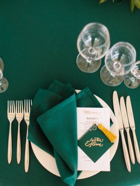Wedding banquet table setting in emerald green. On a white plate are a restaurant menu and a folded napkin and a green sign for the groom's seat.