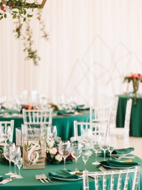 Wedding banquet table decor in emerald green. On the table is a green napkin on a plate, wine glasses, cutlery, flowers, and table number 