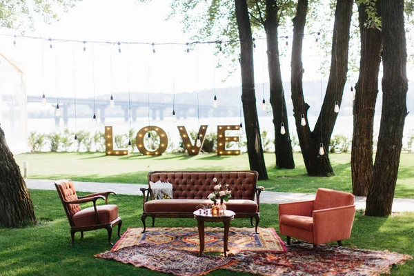 Outdoor party decor: a vintage wooden table with flowers in vases in the center, sofas next to it, and carpets on the lawn. In the background are glowing letters \