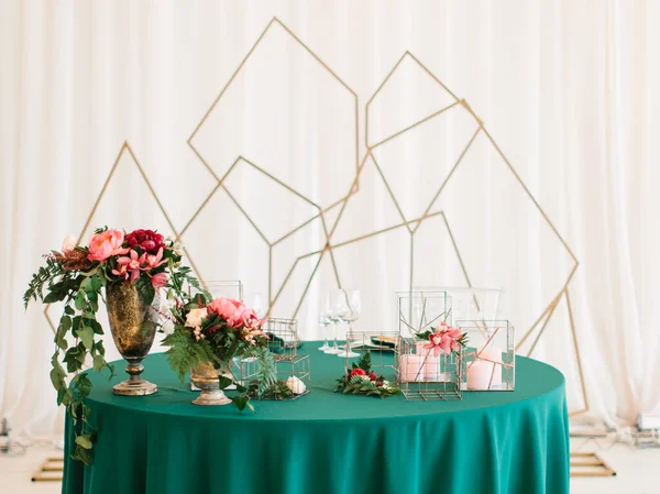 Banquet table decor. On an emerald, green tablecloth is a copper vase with pink and red peonies, flowers in geometric metal stands, and candles in glass boxes. In the background is a golden geometric