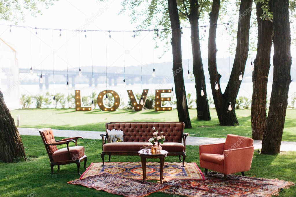 Outdoor party decor: a vintage wooden table with flowers in vases in the center, sofas next to it, and carpets on the lawn. In the background are glowing letters 