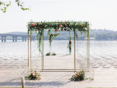 A golden square wedding arch decorated with white, pink, and red peonies stands on a wooden pier. Below are bouquets of flowers and decorative golden cubes. In the background, there is a river.