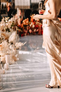 A girl in a beige silk dress that develops in the wind stands and holds a glass of champagne. In the background is a glossy white floor and a decor of flowers and candles.