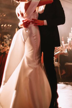 The newlyweds dance at the wedding in the warm light of the spotlight. A girl in a white wedding dress with a long train with an open back, her husband kisses and hugs her.