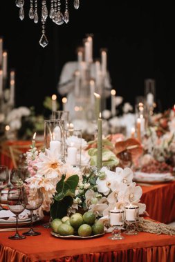 Wedding banquet table decor with an orange tablecloth. In the foreground are lime fruits on a tray, white candles in candlesticks, and black glasses. In the background are white flowers, candles, green leave