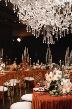 Wedding decor of the banquet table in the restaurant in orange and white colors. Orange tablecloth, copper chairs, white flowers, candles, lime. Crystal chandeliers hang over the table.