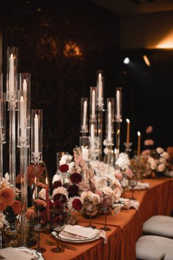Floral decoration of the wedding banquet table in red-orange color. Orange chairs, white flowers, red anthurium, dahlias, burning candles, crockery, cutlery, and beige chairs.