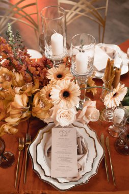 A paper menu template that lies on a plate with a silk napkin. Orange tablecloth, gerberas, daffodils, white candles.