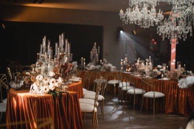 Wedding decor of the banquet table in the restaurant in orange and white colors. Orange tablecloth, copper chairs, white flowers, candles, lime. Crystal chandeliers hang over the table.