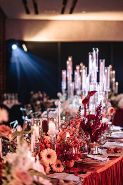 Wedding table decor on an orange tablecloth with white flowers, white china, and black glasses. White tall candles burn in glass candlesticks.