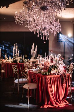 Wedding table decor on an orange tablecloth with white flowers, white china, and black glasses. White tall candles burn in glass candlesticks.