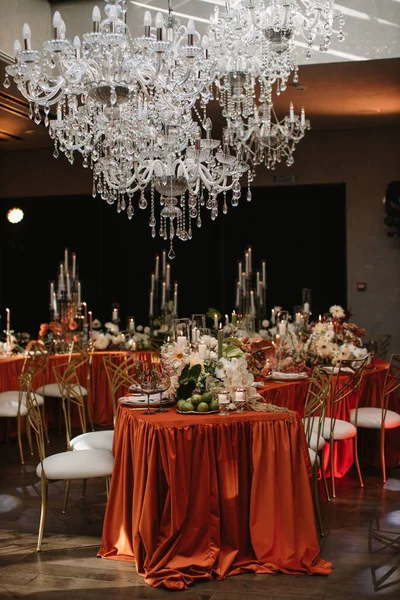 Wedding decor of the banquet table in the restaurant in orange and white colors. Orange tablecloth, copper chairs, white flowers, candles, lime. Crystal chandeliers hang over the table.
