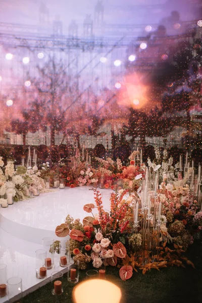 A round wedding photo zone with a white floor and garlands hanging from above. Around the flower arrangements in white and red colors, white candles in glass cases, and red candles.