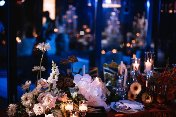 Evening decor of a banquet table in a dark room. Bouquets of flowers, crockery, and wine glasses are illuminated by white candles.