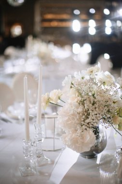 Wedding decor of a banquet table of snow-white color. White roses, peonies, gypsophila, white candles in glass candlesticks.