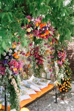 Wrought iron garden arch in an Italian garden with a bench, an orange cushion, and pillows depicting tangerines. The arch is entwined with bright multi-colored flowers, in the background is a tangerine