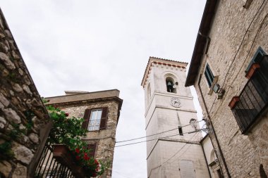 Narrow Italian street view from the bottom up to the upper floors of stone houses and a white rectangular chapel with bells.