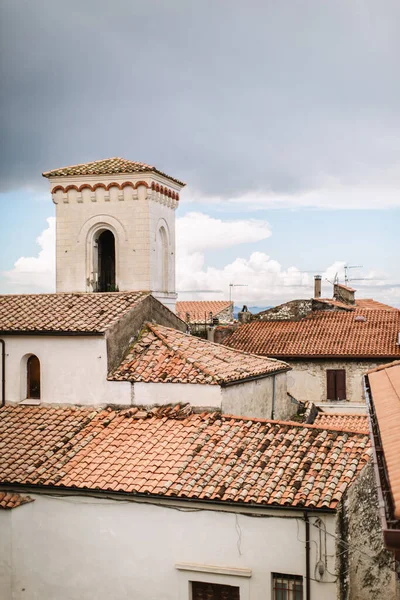 Antique pitched roof with old red tiles on an Italian house. In the background are a white tower, a red roof, and brick chimneys.