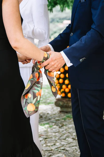 A man in a dark blue suit holds the hands of a woman in a black dress close-up. A woman has a brown silk scarf with a print of orange tangerines tied around her arm.