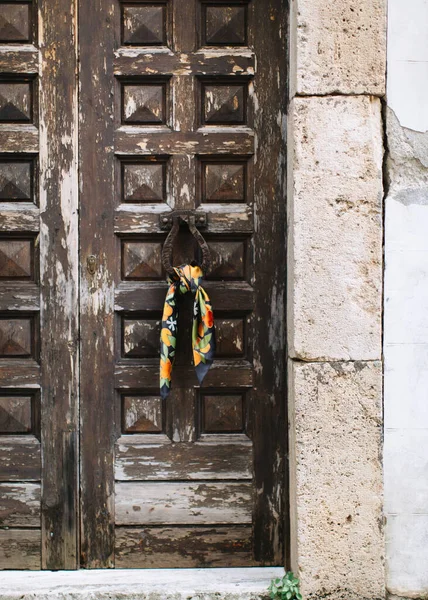 A woman\'s silk scarf with tangerines is tied to a carved door knocker, an old door with peeling paint. To the right of the door is a stone wall.