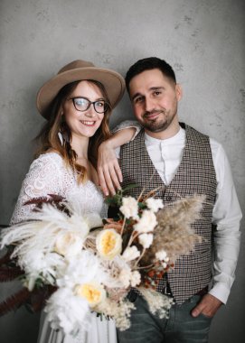 Boho tarzı giyinmiş mutlu gelin ve damat gri beton duvarın yanında duruyor. Şapkalı ve tüylü gelin elinde şakayık ve dikenli buket ve gülücüklerle.