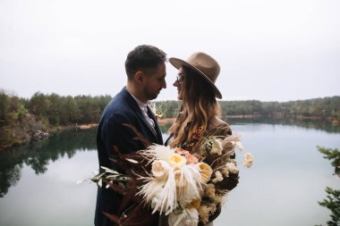 The bride and groom stand on a stone near the lake in the forest and kiss. The bride holds a wedding bouquet in boho style. Wedding in the forest.